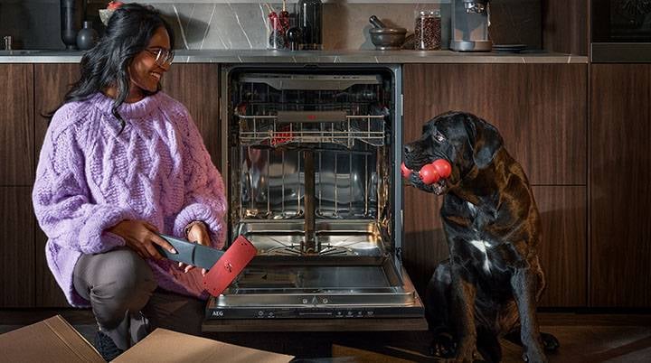 Kvinna packar upp reservdel till diskmaskin medan hon tittar på en svart hund med leksak i munnen