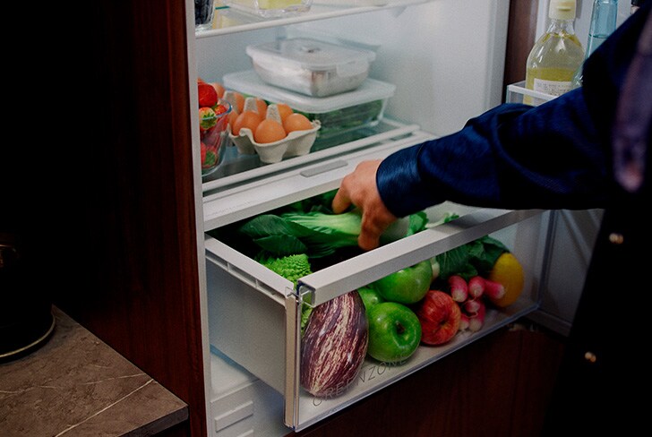 Öppet kylskåp med frukt och grönsaker i grönsakslåda och person som organiserar innehållet.