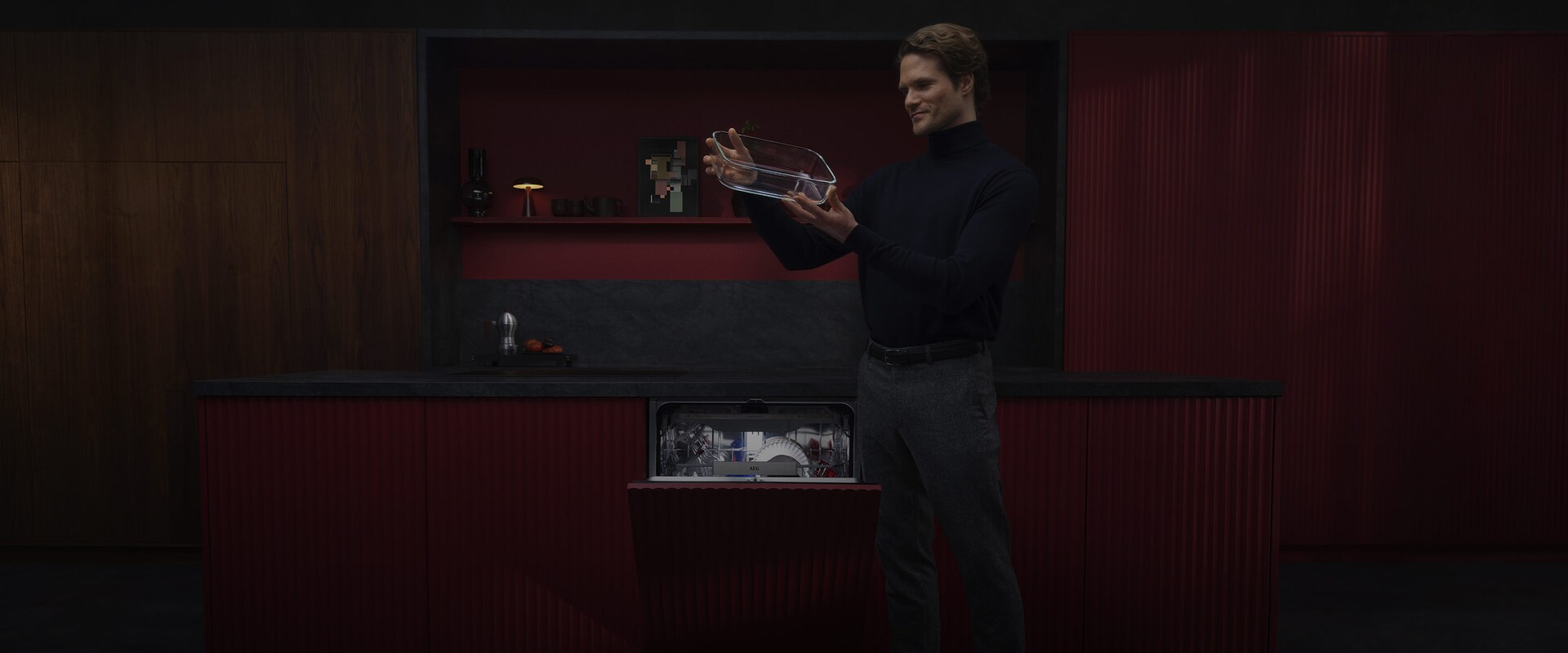 En man i svart polotröja inspekterar en ren ugnsform i glas framför en öppen AEG-diskmaskin i ett djuprött kök.