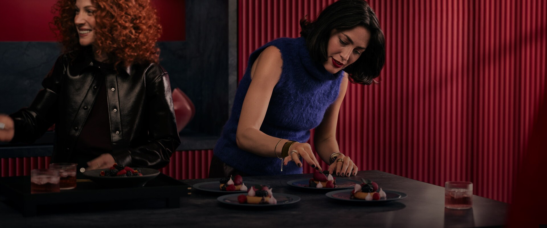 Två kvinnor förbereder desserter på en mörk köksö framför en röd ribbad väggpanel.